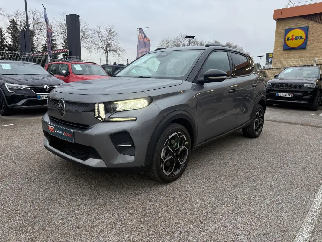 Citroën C3 gris vue 3/4 avant droit, phares allumés, jantes alliage noires et barres de toit visibles.