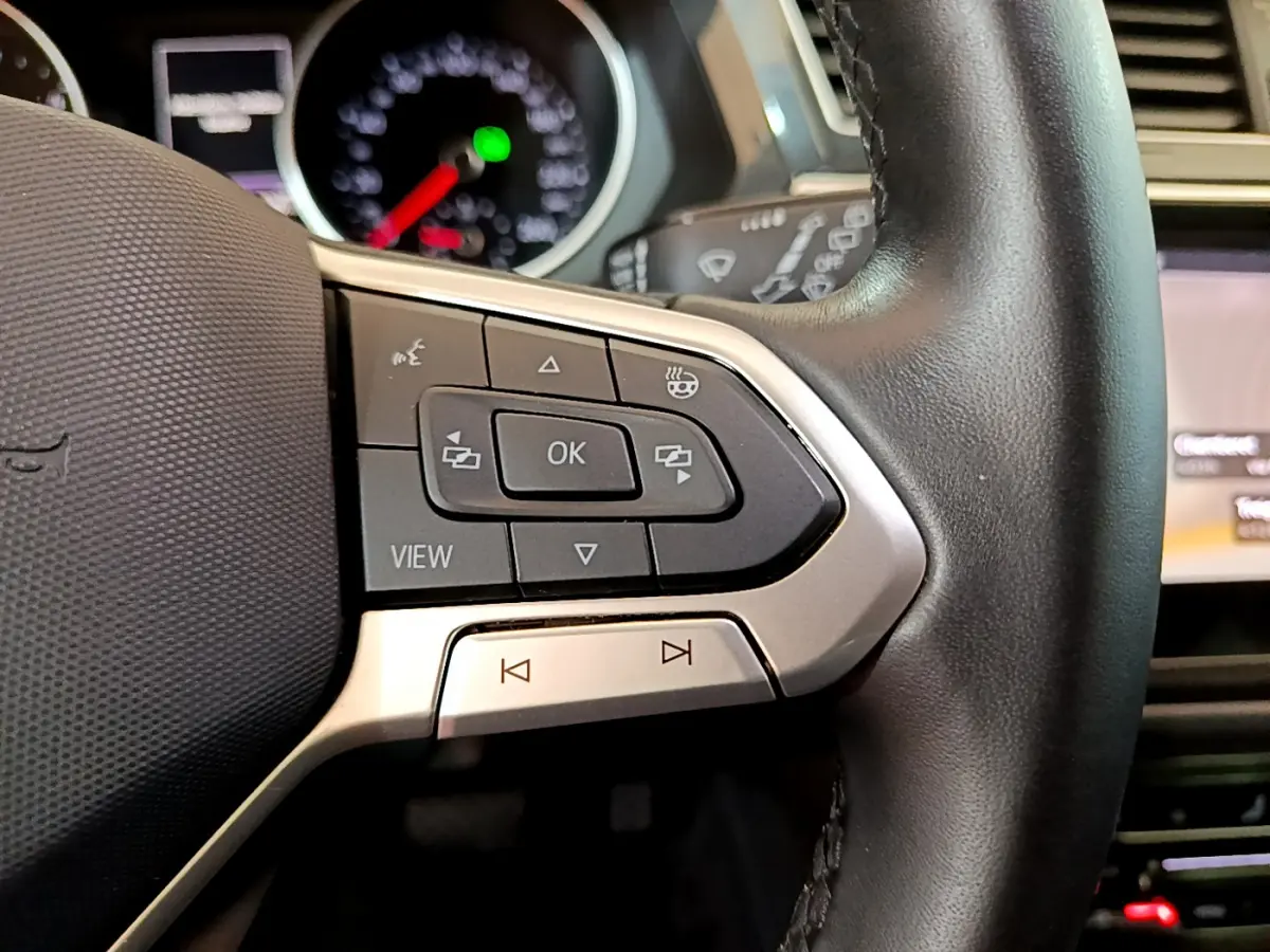 Gros plan sur les commandes gauche du volant cuir du Volkswagen Tiguan gris clair, avec tableau de bord flou en arrière-plan.