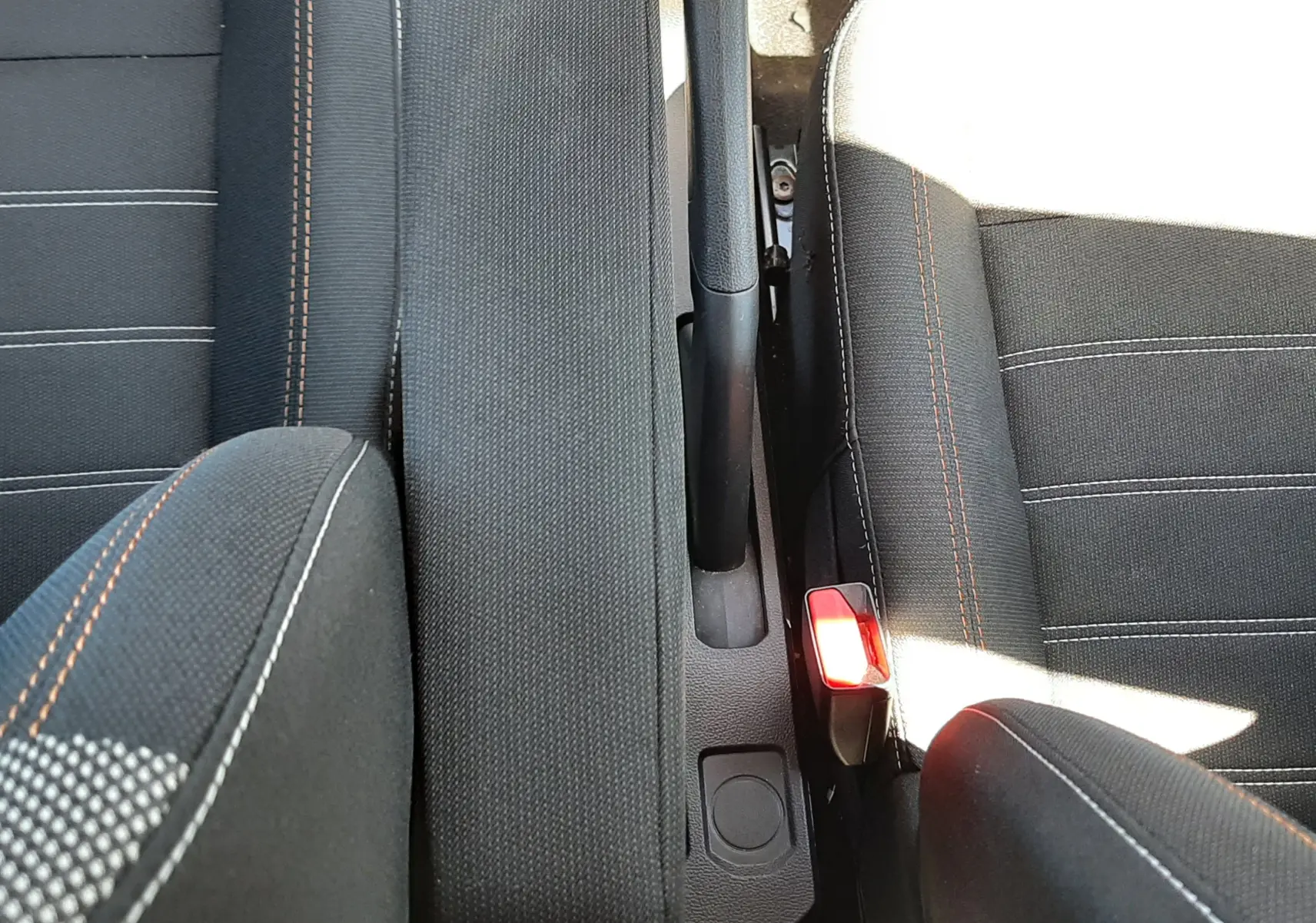 Vue plongeante sur l'accoudoir central noir et le frein à main entre les sièges avant gris foncé avec surpiqûres orange de la Dacia Sandero Stepway.