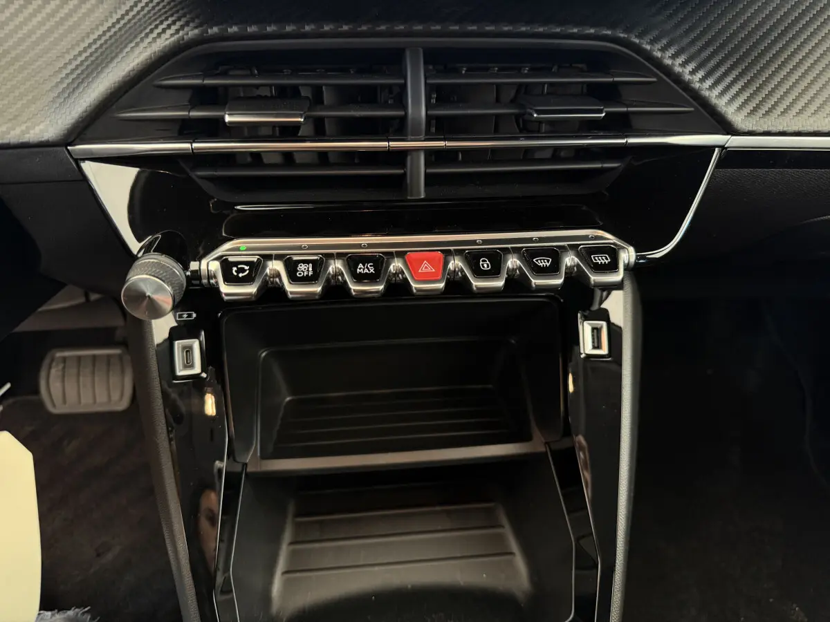 Gros plan sur la console centrale noire d'une Peugeot 208 2021, avec boutons de commande et prises USB visibles.