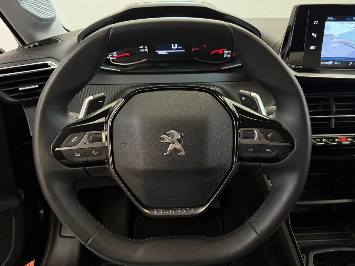 Vue rapprochée du volant cuir noir de la Peugeot 208 2021 avec commandes multifonctions et tableau de bord numérique.