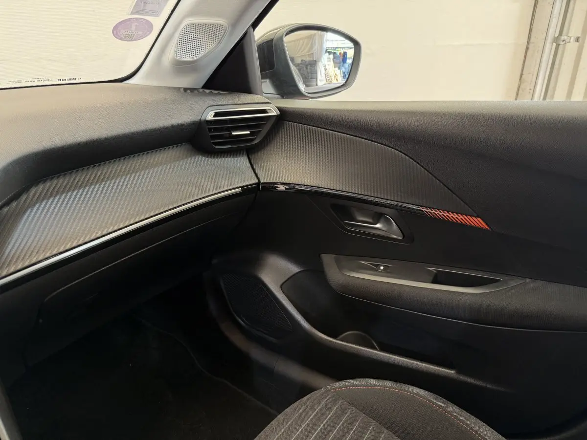 Intérieur côté passager de la Peugeot 208 noire, avec garniture carbone et détail rouge sur la porte.