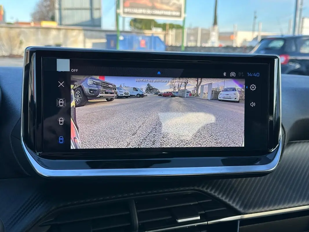 Écran tactile intérieur montrant la caméra de recul d'une Peugeot 208 blanche, vue arrière avec route et véhicules.