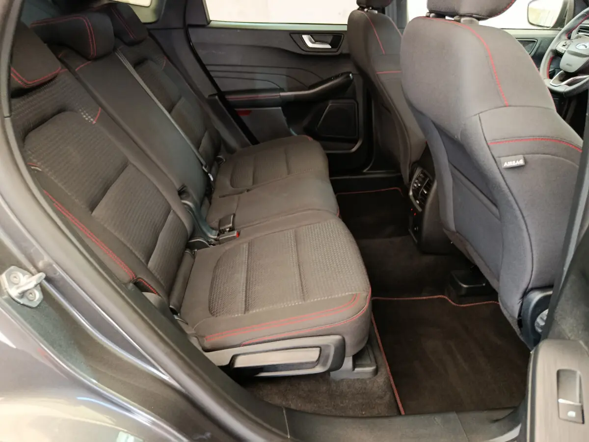 Vue intérieure arrière côté gauche du Ford Kuga 2024 gris clair, sièges tissu noir avec surpiqûres rouges et tapis assortis.
