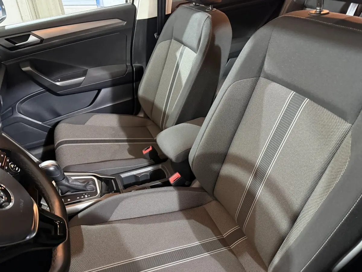 Vue intérieure côté conducteur du Volkswagen T-Roc 1.5 TSI blanc, sièges tissu gris avec bandes claires et console centrale visible.