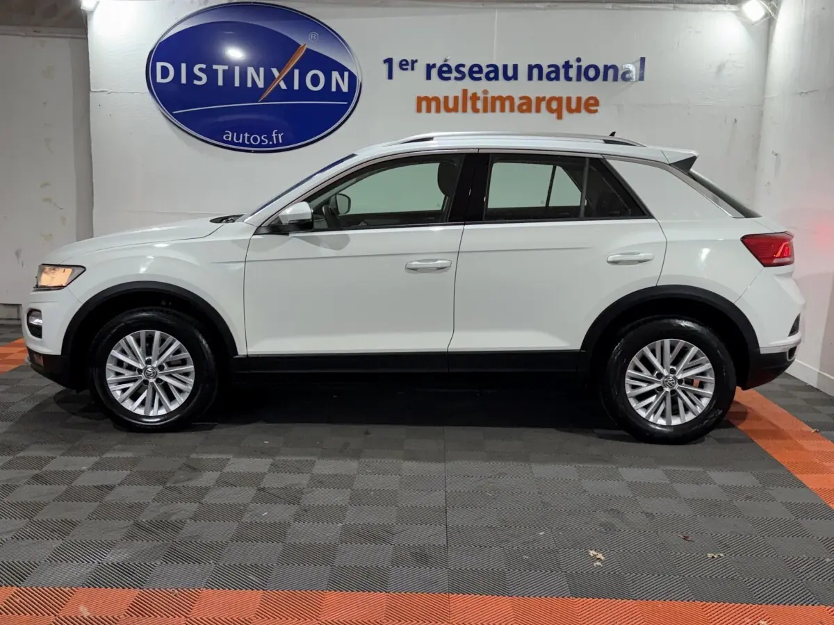 Profil côté gauche d'un Volkswagen T-Roc blanc 2020 avec jantes alliage et barres de toit noires en intérieur showroom.