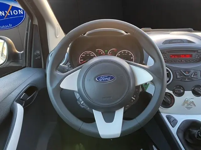 Vue intérieure centrée sur le volant gris et blanc d'une Ford KA 1.2 69 Trend blanche de 2015 avec tableau de bord et console visibles.