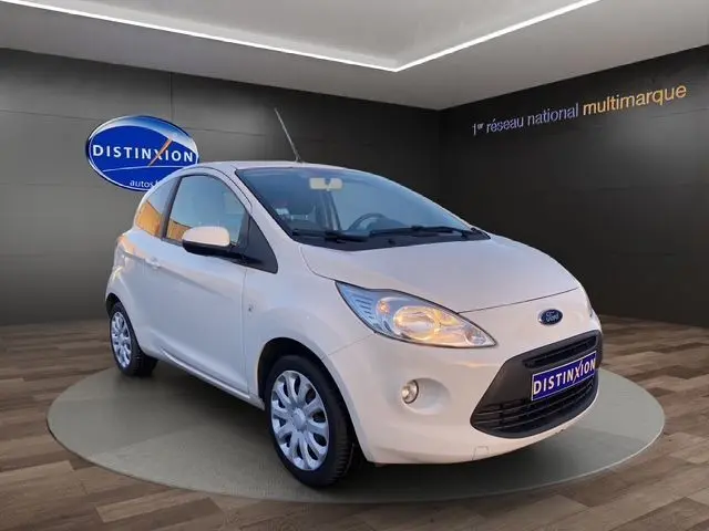 Vue 3/4 avant d'une Ford KA blanche de 2015, compacte avec phares allumés dans un showroom.