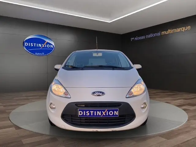 Vue frontale d'une Ford KA blanche de 2015 avec phares allumés, sur fond intérieur showroom Distinxion.