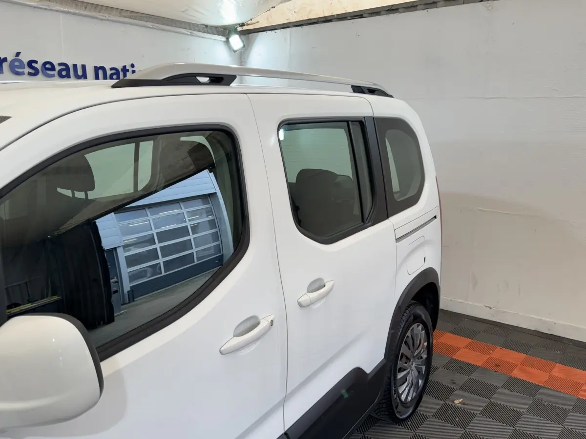 Vue latérale droite d’un Peugeot Rifter BlueHDi 100 Allure blanc, avec barres de toit et jantes alliage visibles.