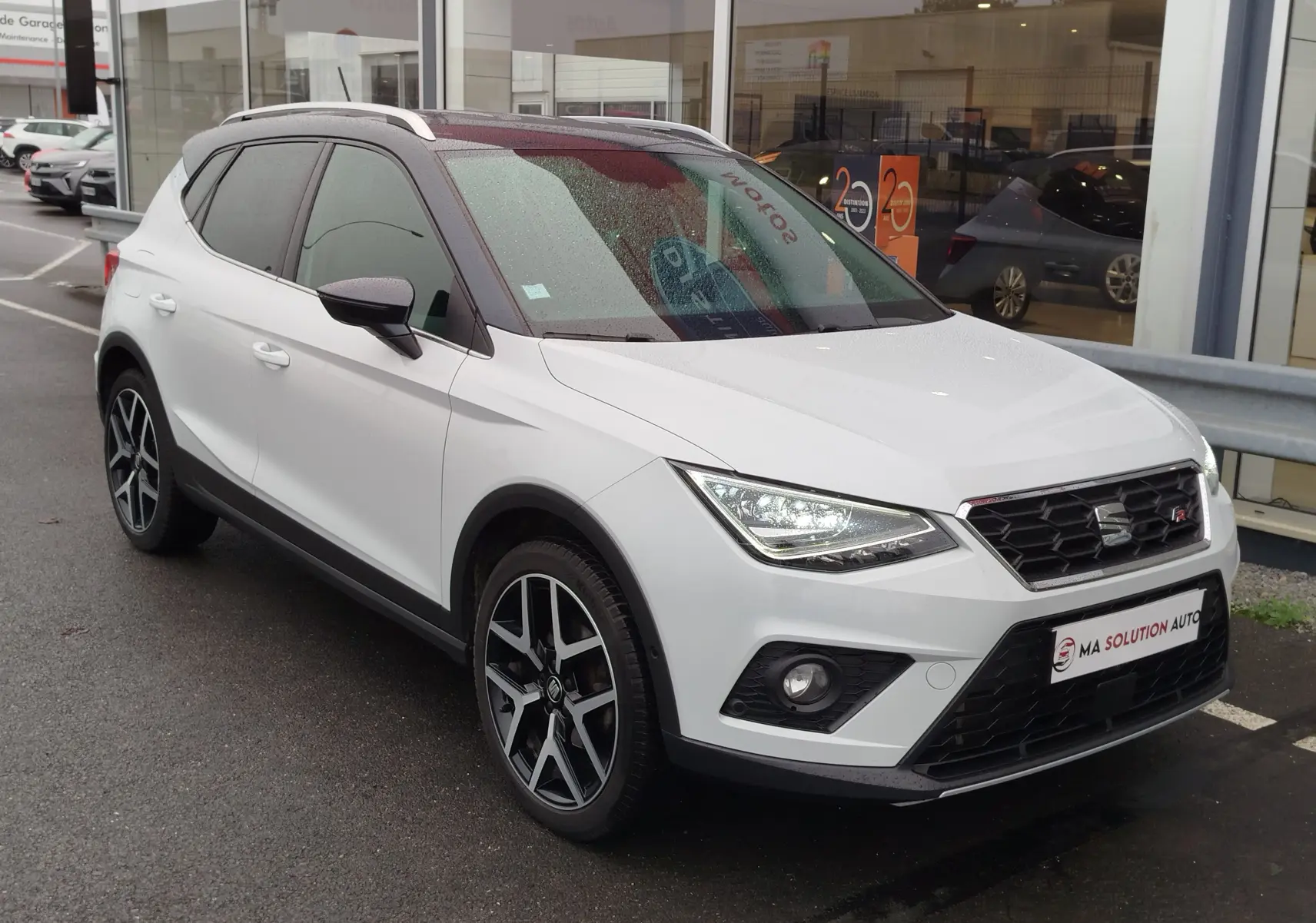 SEAT Arona blanc avec toit gris magnétique, vue 3/4 avant droit, feux allumés et jantes alliage noires.