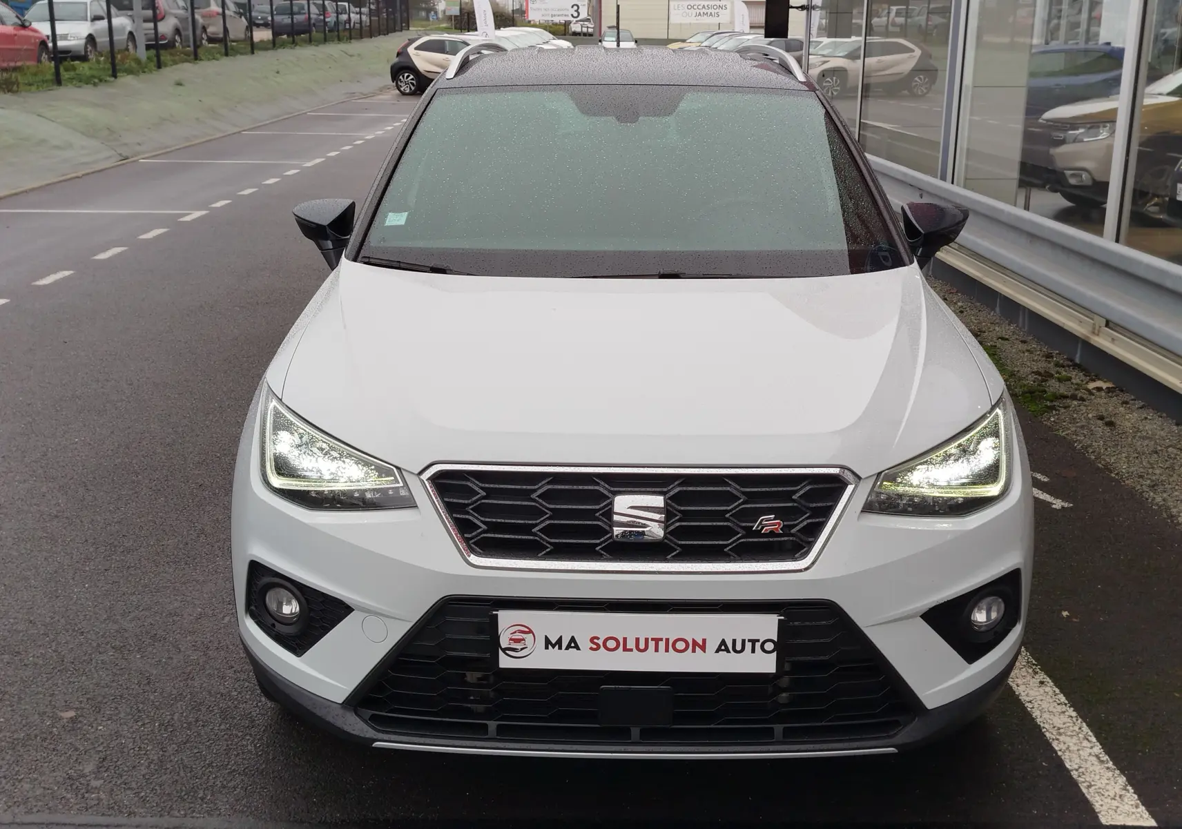 Vue frontale d'un SEAT Arona blanc avec toit gris magnétique, phares allumés et calandre noire hexagonale.