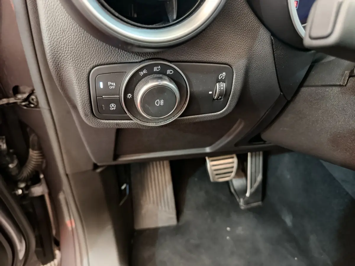 Gros plan sur la commande des phares et les pédales côté gauche de l'Alfa Romeo Stelvio gris foncé 2024 Sprint.