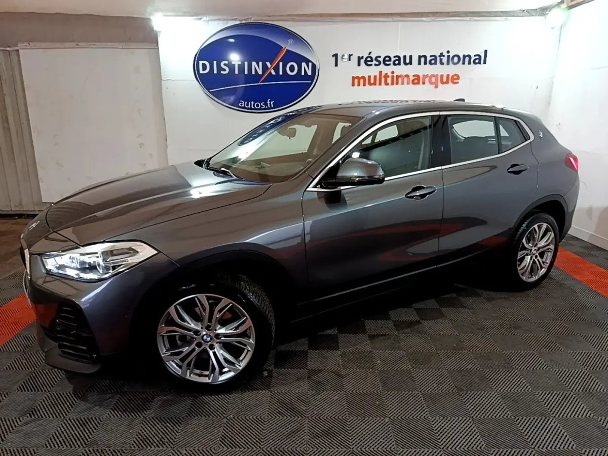 BMW X2 sDrive18i gris foncé vue de profil côté gauche, avec jantes alliage et phares allumés dans un showroom.