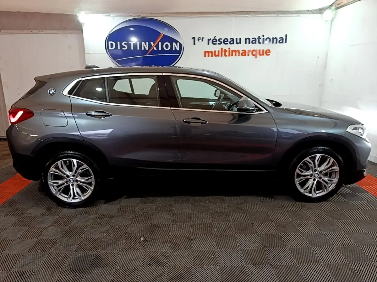 BMW X2 sDrive18i Lounge gris foncé vue de profil côté gauche, avec jantes alliage et feux arrière allumés.
