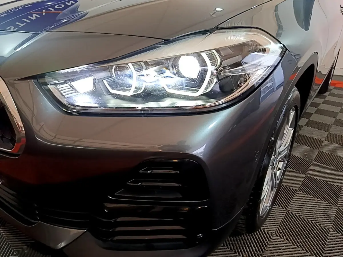 Gros plan sur le phare avant droit allumé d’un BMW X2 gris foncé, avec partie de la roue visible.