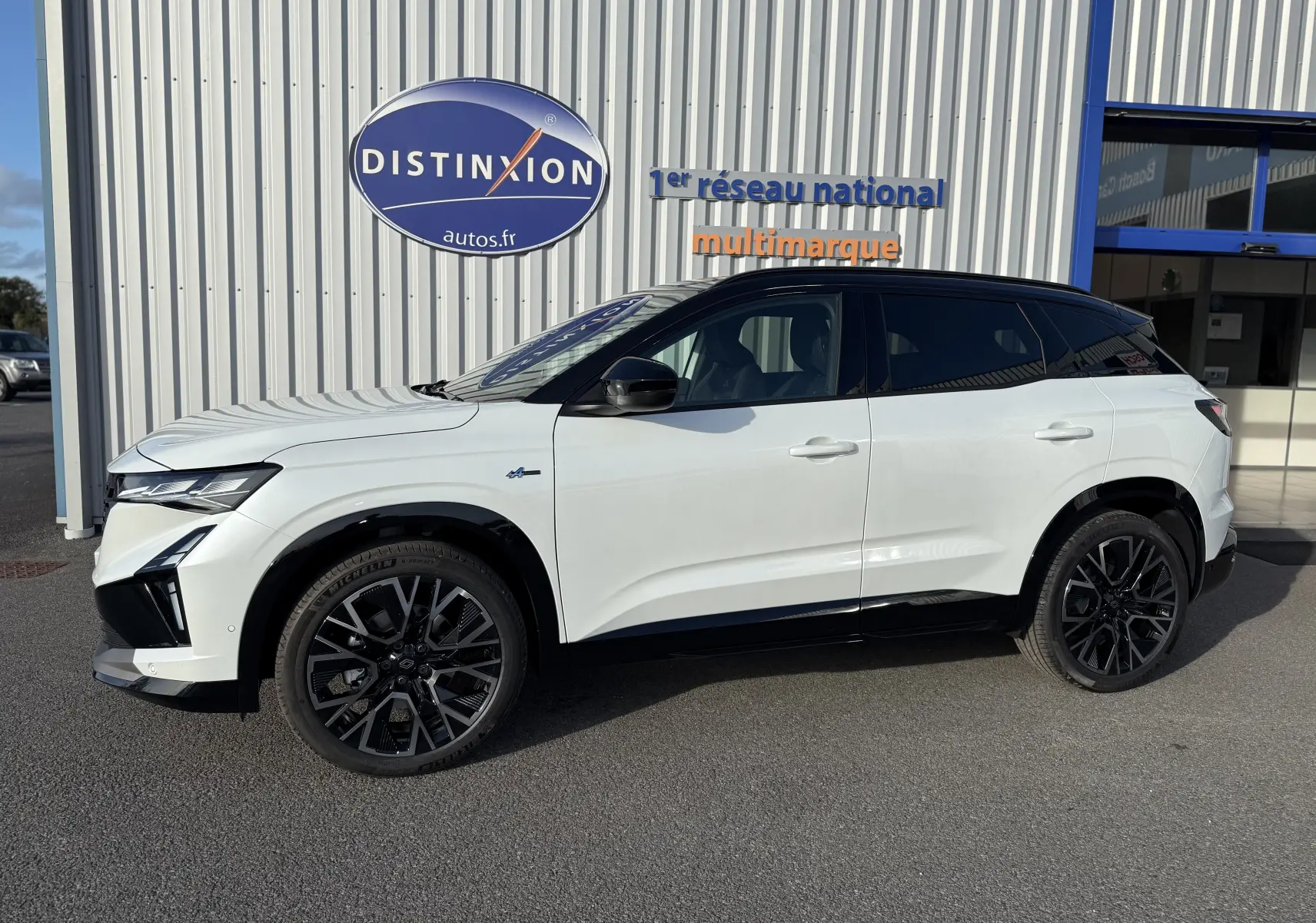 Profil côté gauche du Renault Austral blanc nacré avec toit noir et jantes noires, stationné devant un bâtiment commercial.