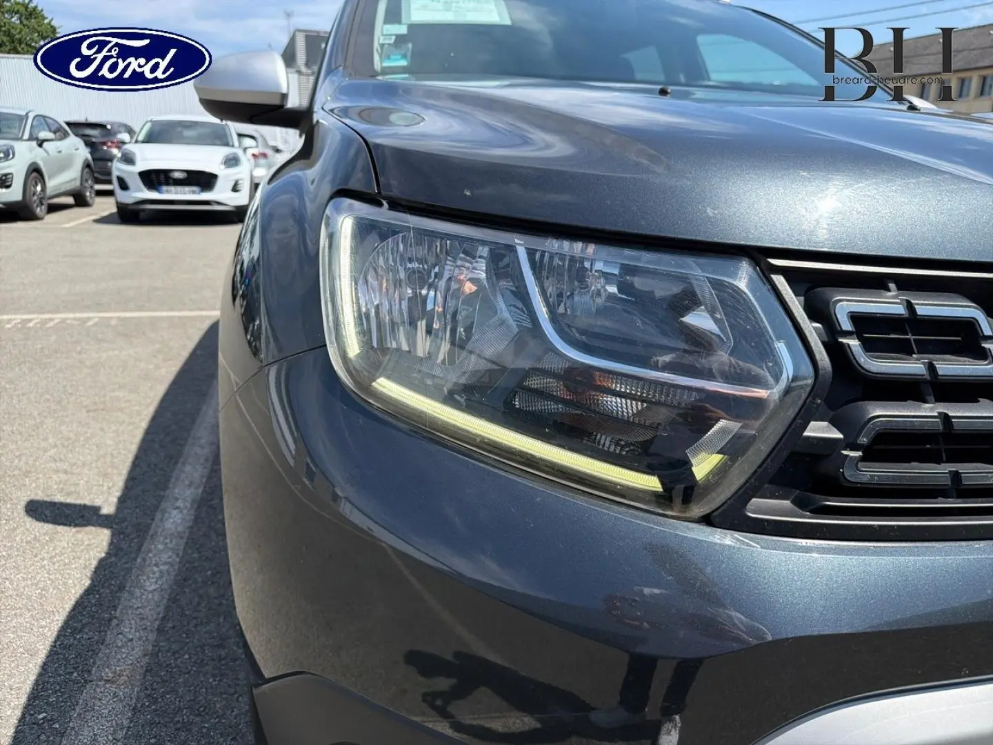 Gros plan sur le phare avant gauche et la calandre d’un Dacia Duster gris Comète métallisé, vue de face.