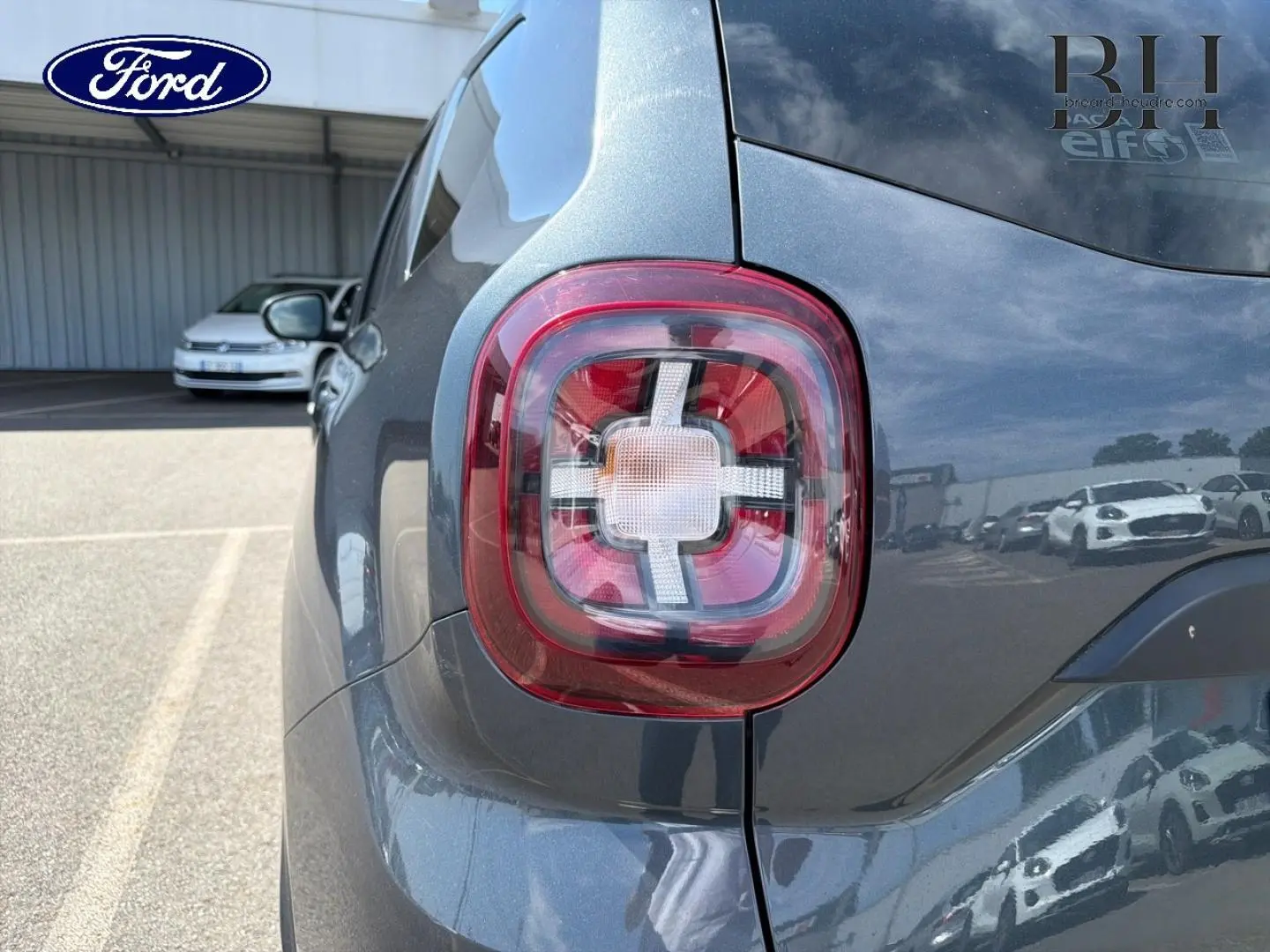 Gros plan sur le feu arrière droit du Dacia Duster gris Comète métallisé, avec reflet du ciel et voitures alentour.
