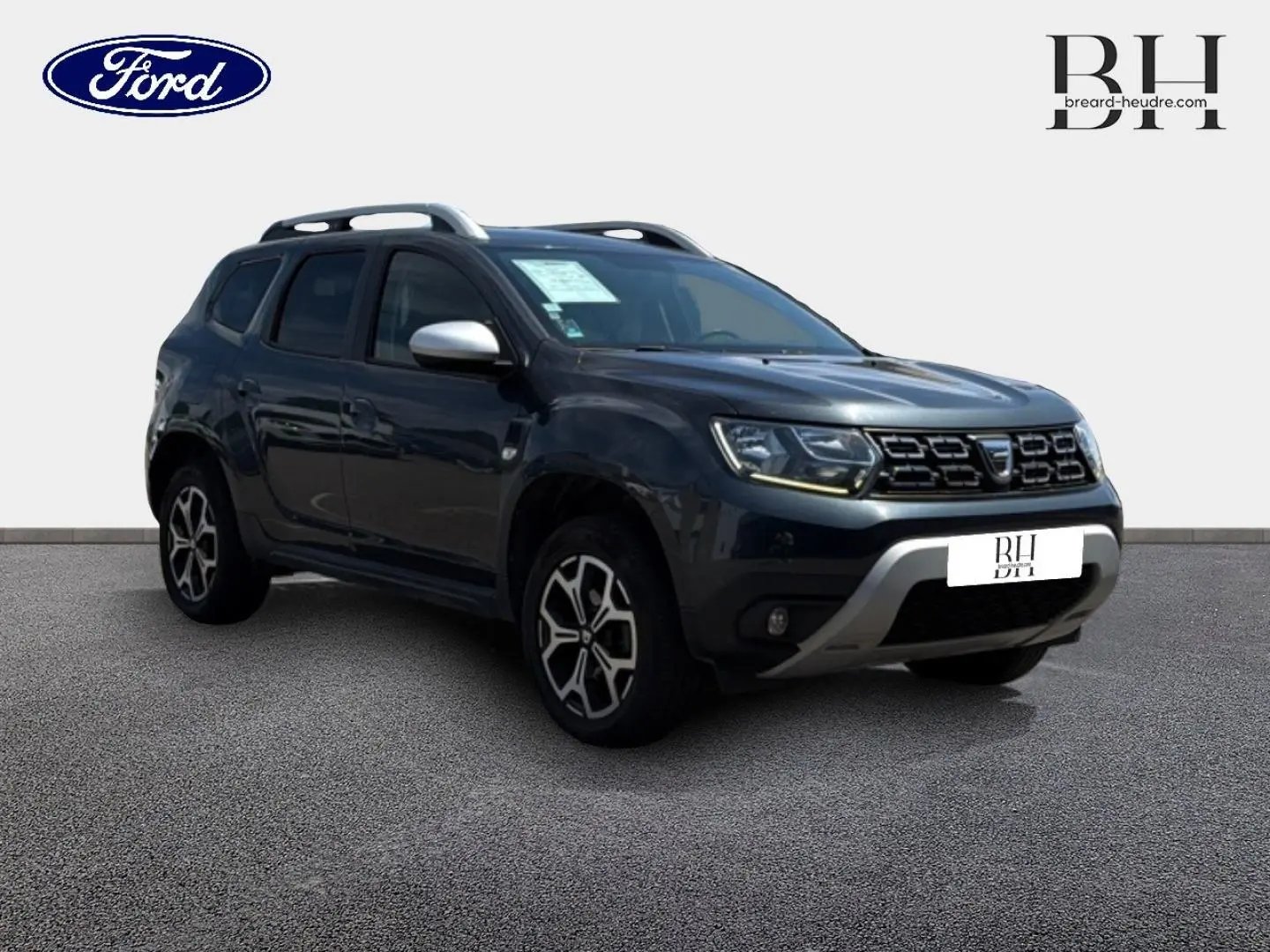 Dacia Duster gris Comète métallisé en 3/4 avant droit, avec barres de toit et jantes alliage visibles.