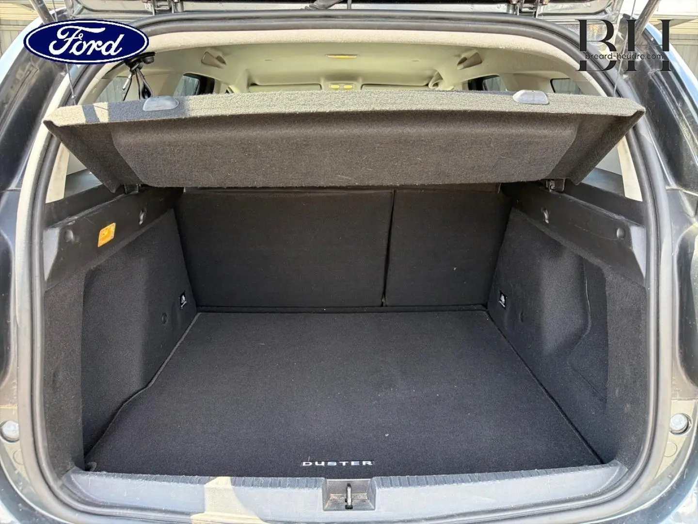 Coffre ouvert vu de l'arrière d'un Dacia Duster gris avec tapis de coffre noir marqué Duster.
