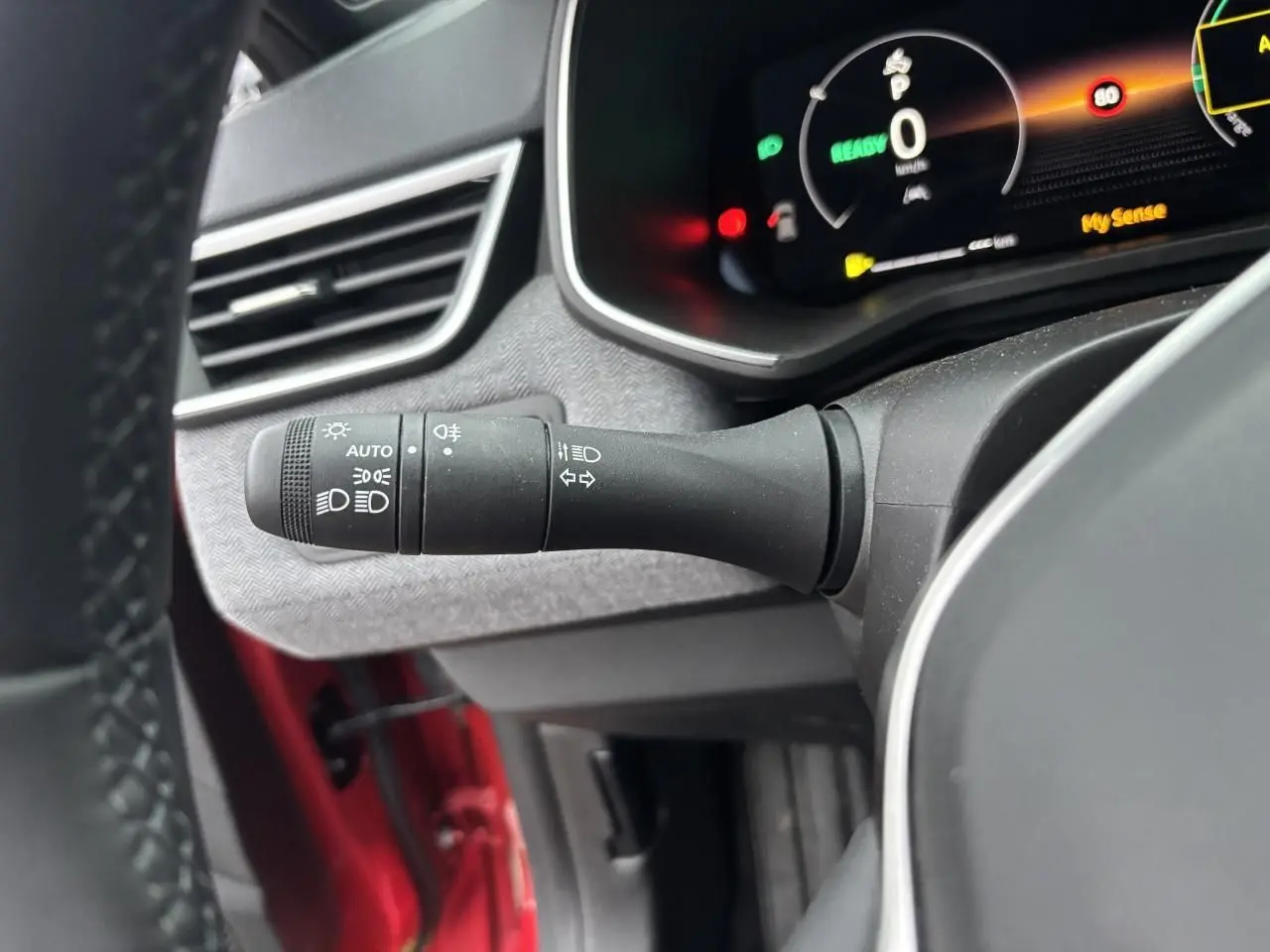Gros plan sur la commande des phares et clignotants côté gauche du volant, intérieur gris et rouge flamme visible.