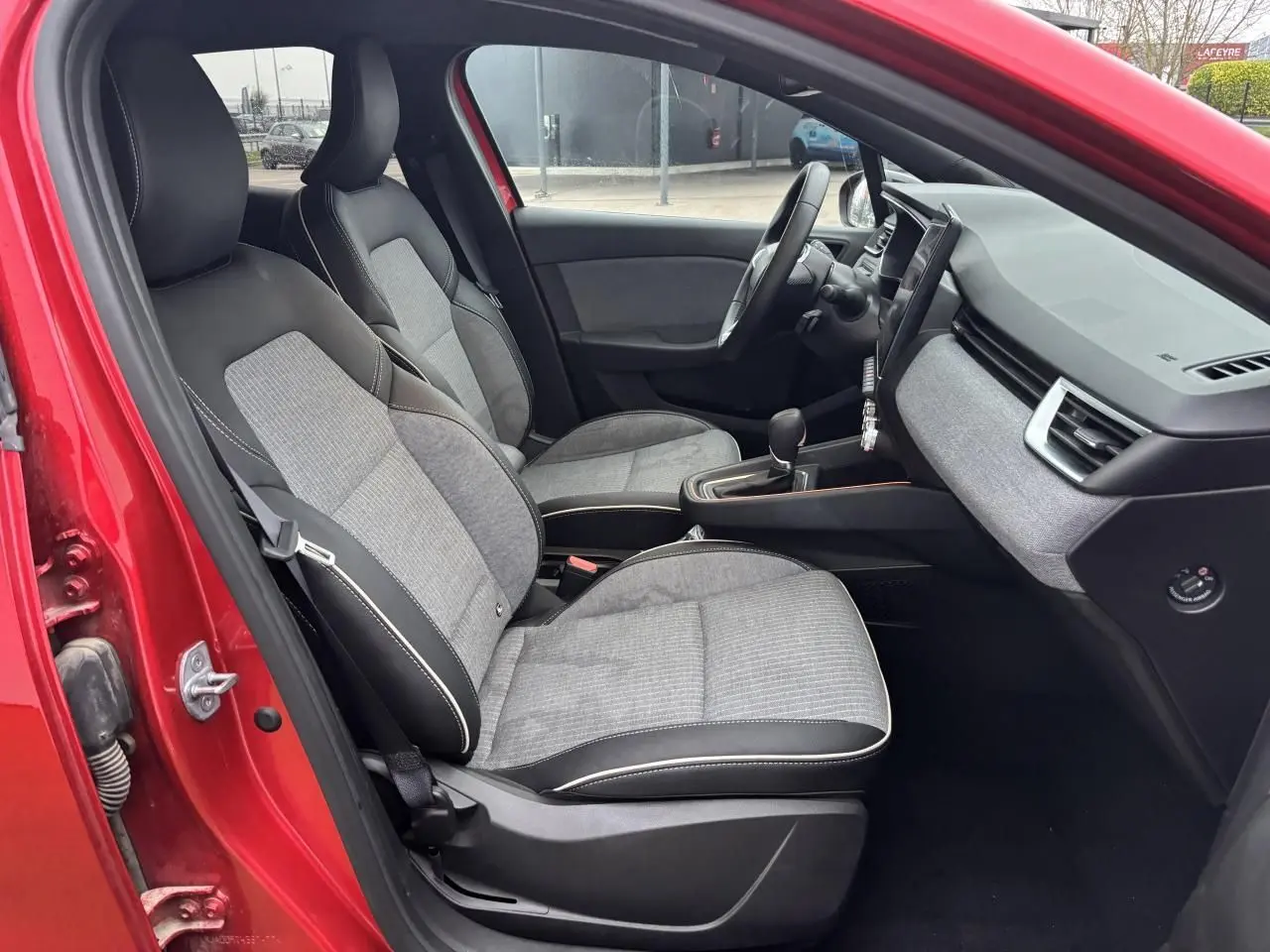 Vue intérieure côté conducteur de la Renault Clio V rouge flamme, montrant sièges gris mélangé et tableau de bord moderne.