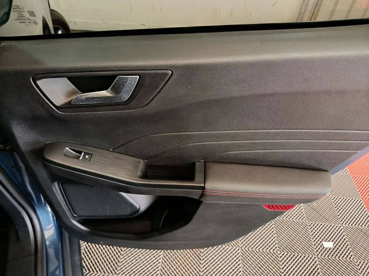 Gros plan sur la porte avant gauche du Ford Kuga bleu 2024, avec poignée argentée et surpiqûres rouges sur l'accoudoir.