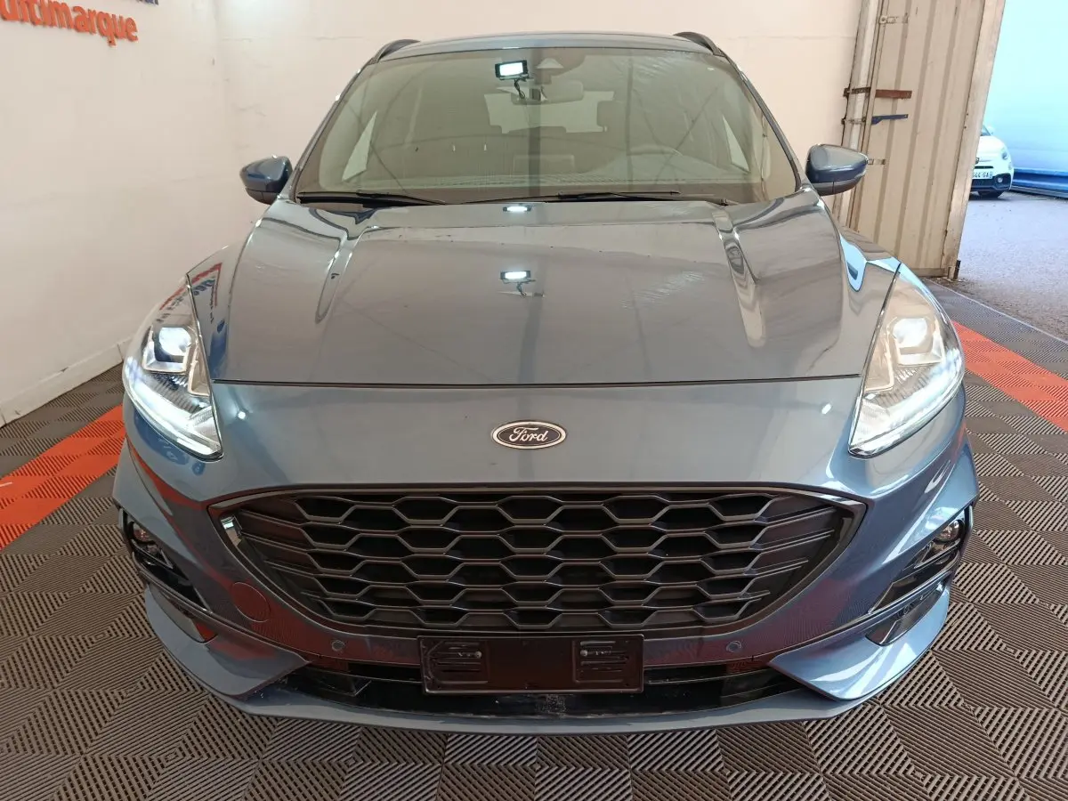 Vue frontale rapprochée du Ford Kuga bleu 2024 avec calandre noire et phares allumés en intérieur.