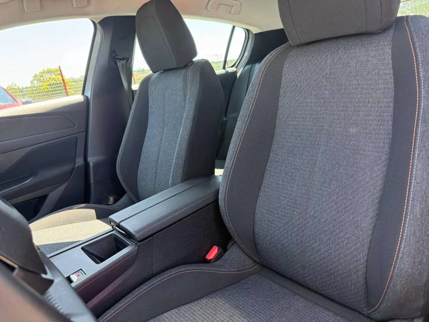 Gros plan sur les sièges avant gris tissu avec surpiqûres orange et accoudoir central de la Peugeot e-308 2025.