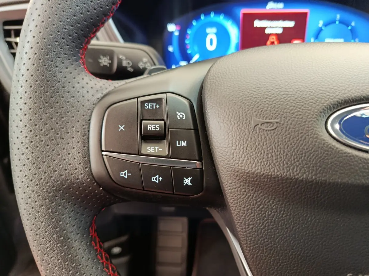 Gros plan sur le volant noir perforé avec surpiqûres rouges du Ford Kuga 2024, affichage numérique bleu en arrière-plan.