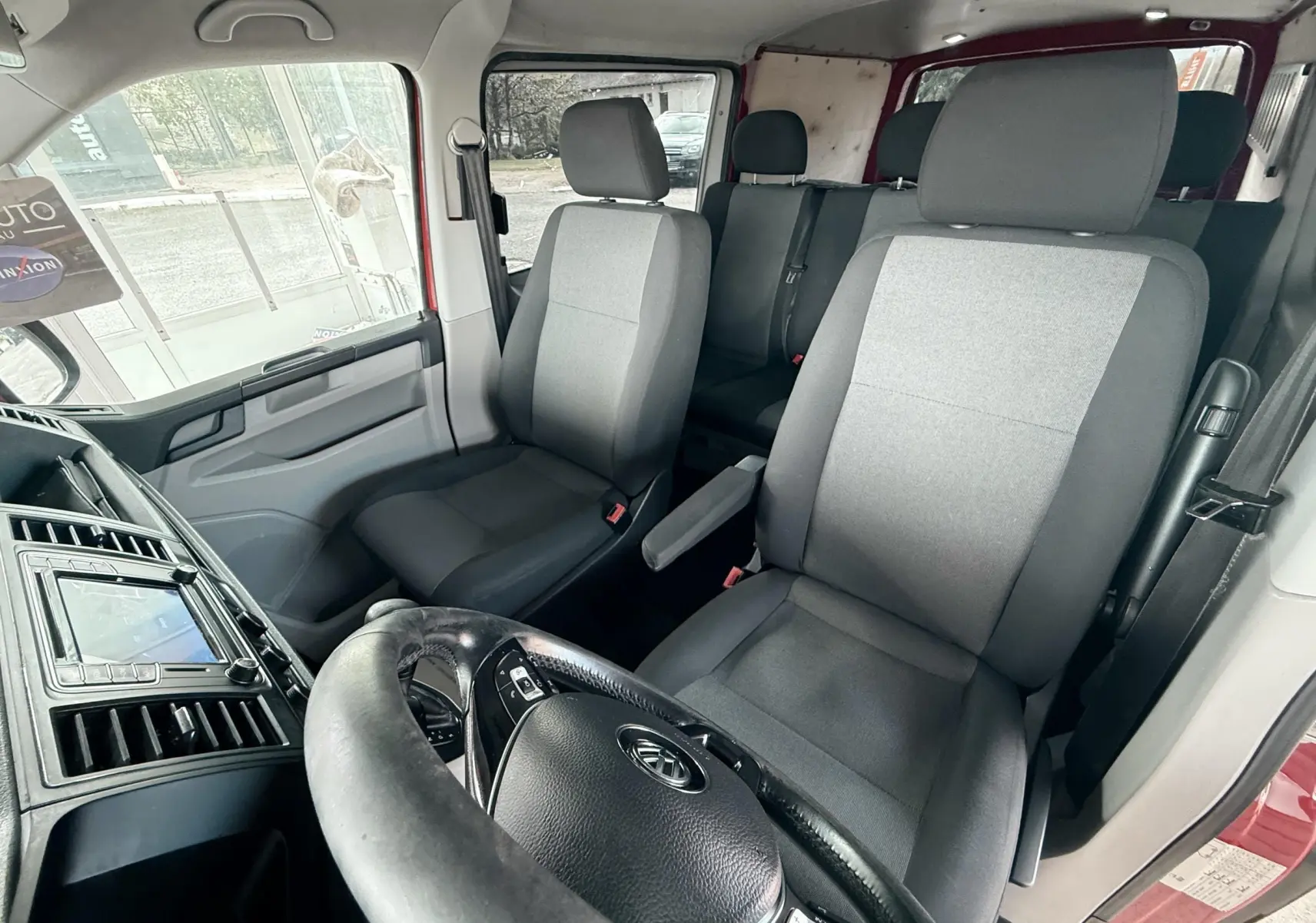 Vue intérieure avant du Volkswagen Transporter rouge, montrant les sièges en tissu gris et le volant noir.