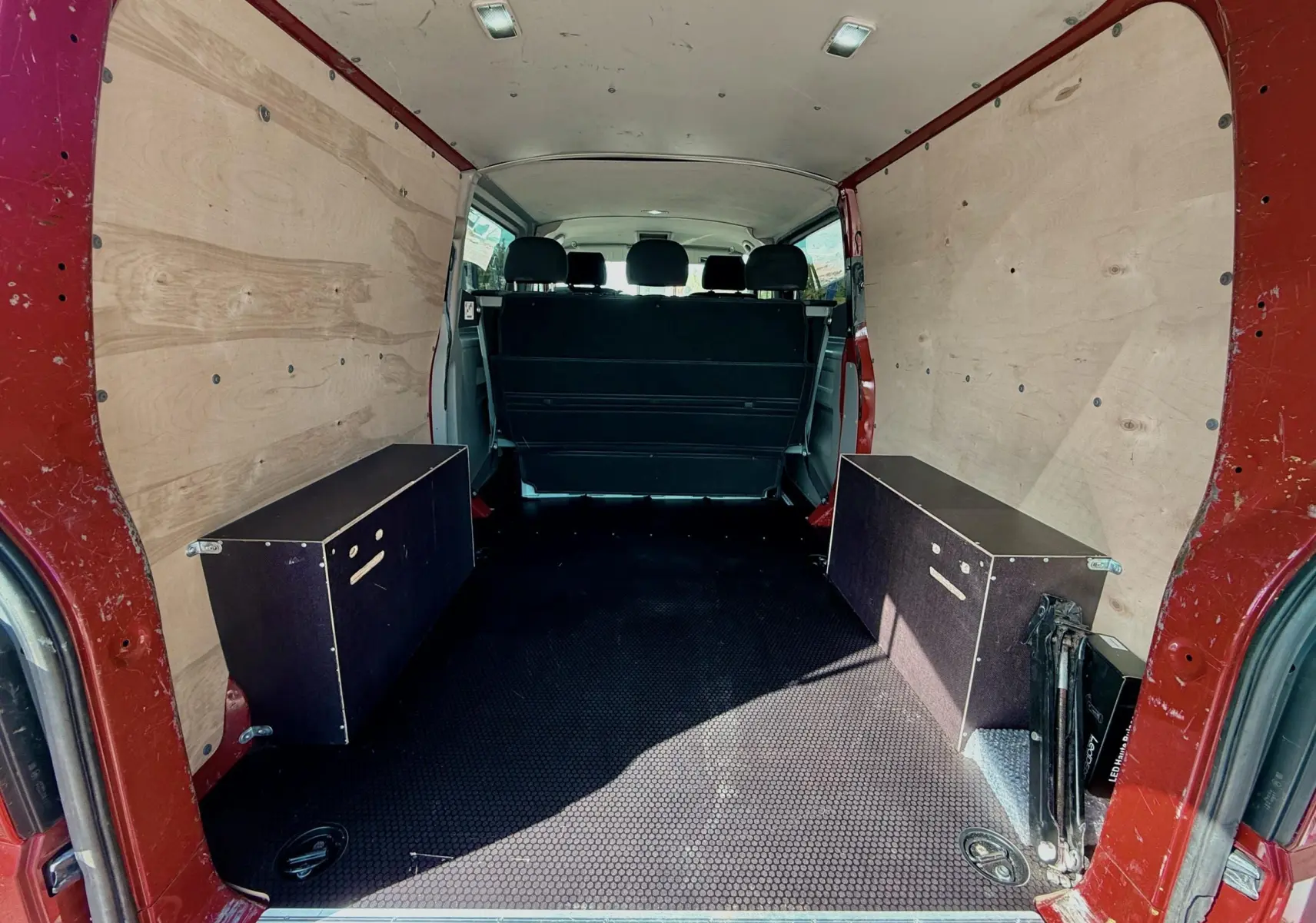 Vue intérieure arrière du Volkswagen Transporter rouge, montrant l'espace de chargement avec panneaux bois et banquette noire.