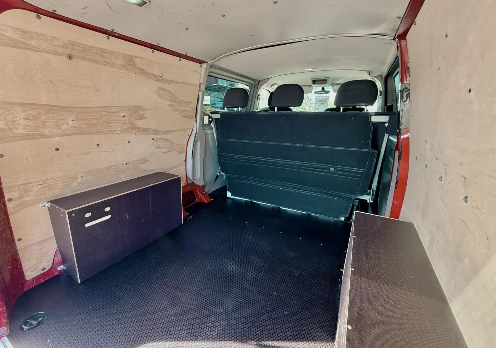 Vue intérieure arrière du Volkswagen Transporter rouge, montrant l’espace de chargement avec banquette et panneaux en bois.