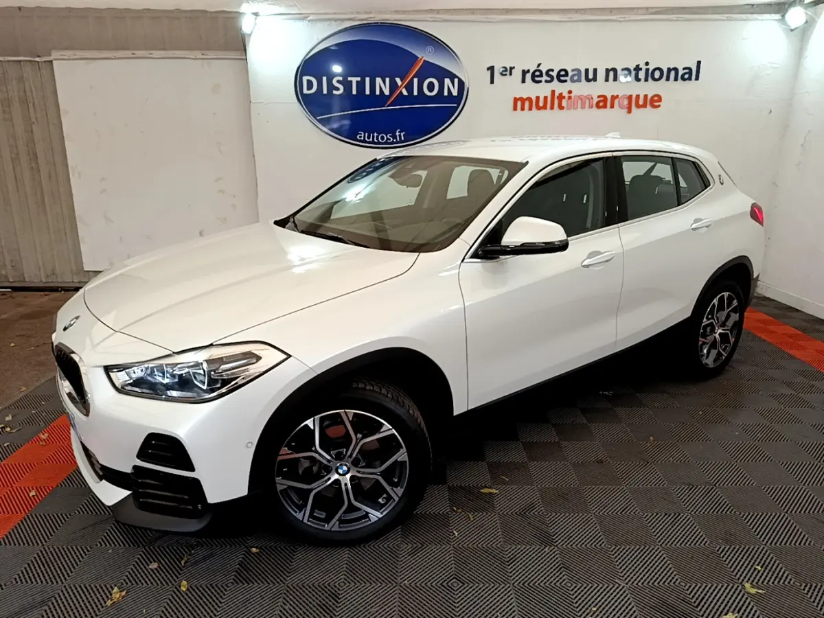 BMW X2 blanc en 3/4 avant droit dans un showroom avec jantes alliage et phares LED allumés.