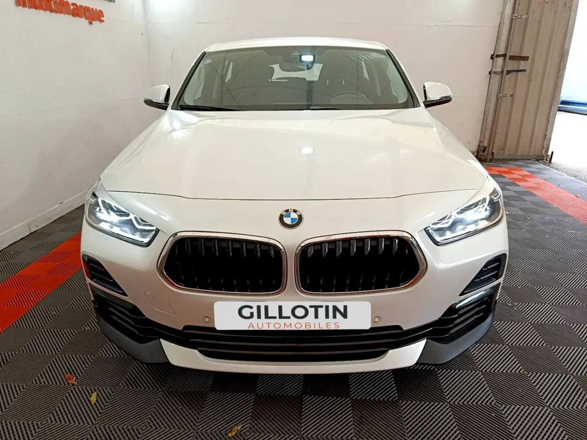 Vue avant d'un BMW X2 blanc 2023 avec calandre double haricot et phares LED allumés dans un garage.