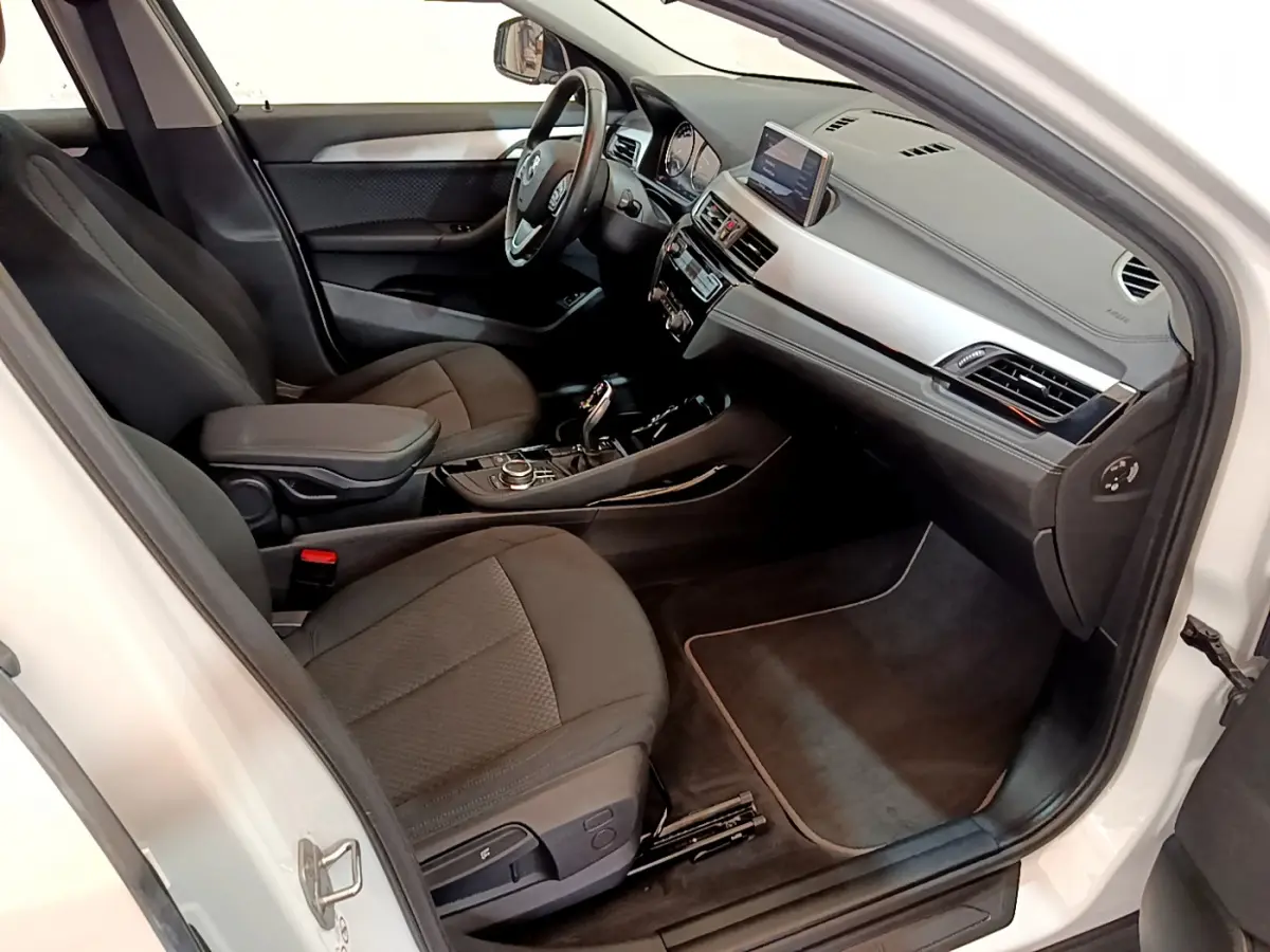 Intérieur BMW X2 sDrive18i blanc 2023 vu côté passager, tableau de bord moderne avec écran central et sièges noirs.