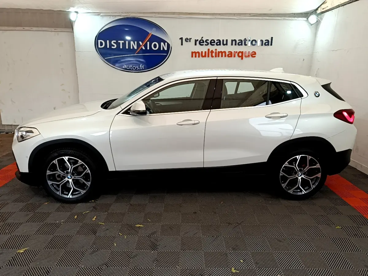 Profil droit d'un BMW X2 blanc 2023 en intérieur, avec jantes alliage et feux arrière LED allumés.