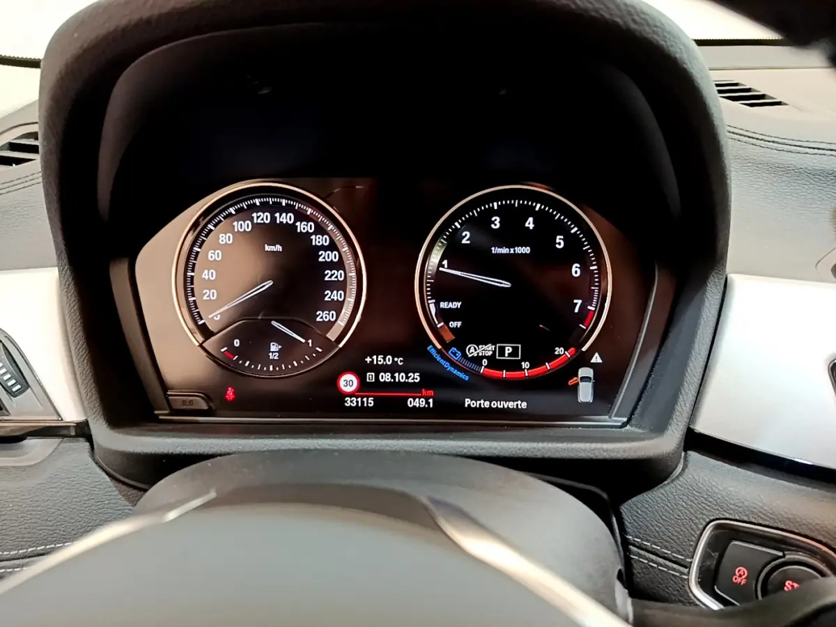 Tableau de bord numérique de BMW X2 blanc 2023, affichage vitesse et compte-tours avec indications de température et porte ouverte.