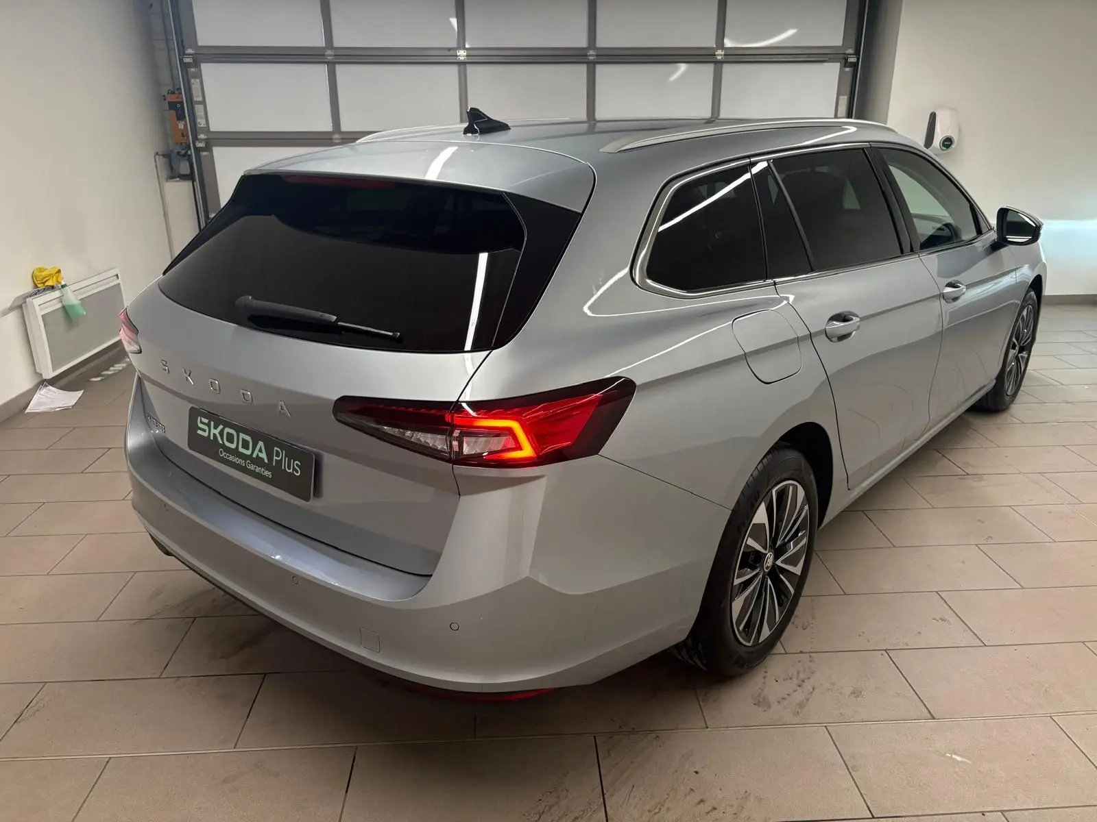 Vue 3/4 arrière droite d'une Skoda Superb Combi gris argent 2025 avec feux arrière allumés dans un garage.