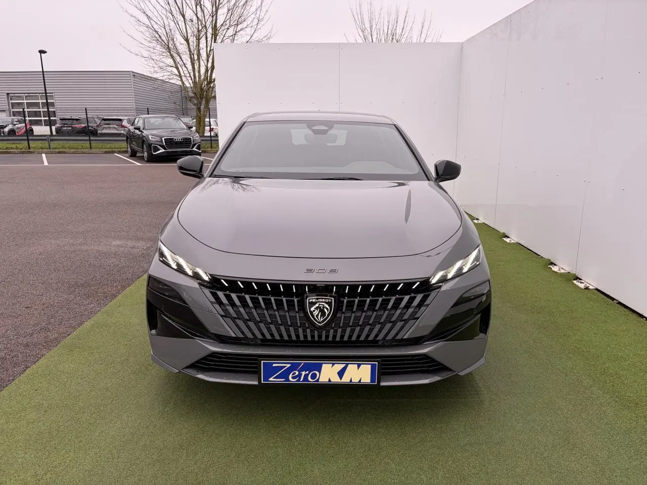 Vue frontale d'une Peugeot 308 III gris selenium avec calandre noire et logo lion lumineux sur fond de parking.