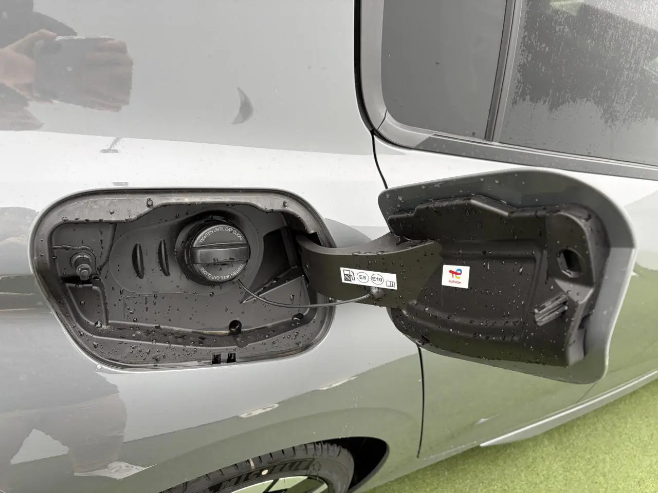 Trappe de carburant ouverte côté gauche d’une Peugeot 308 gris selenium avec bouchon essence visible sous la pluie.