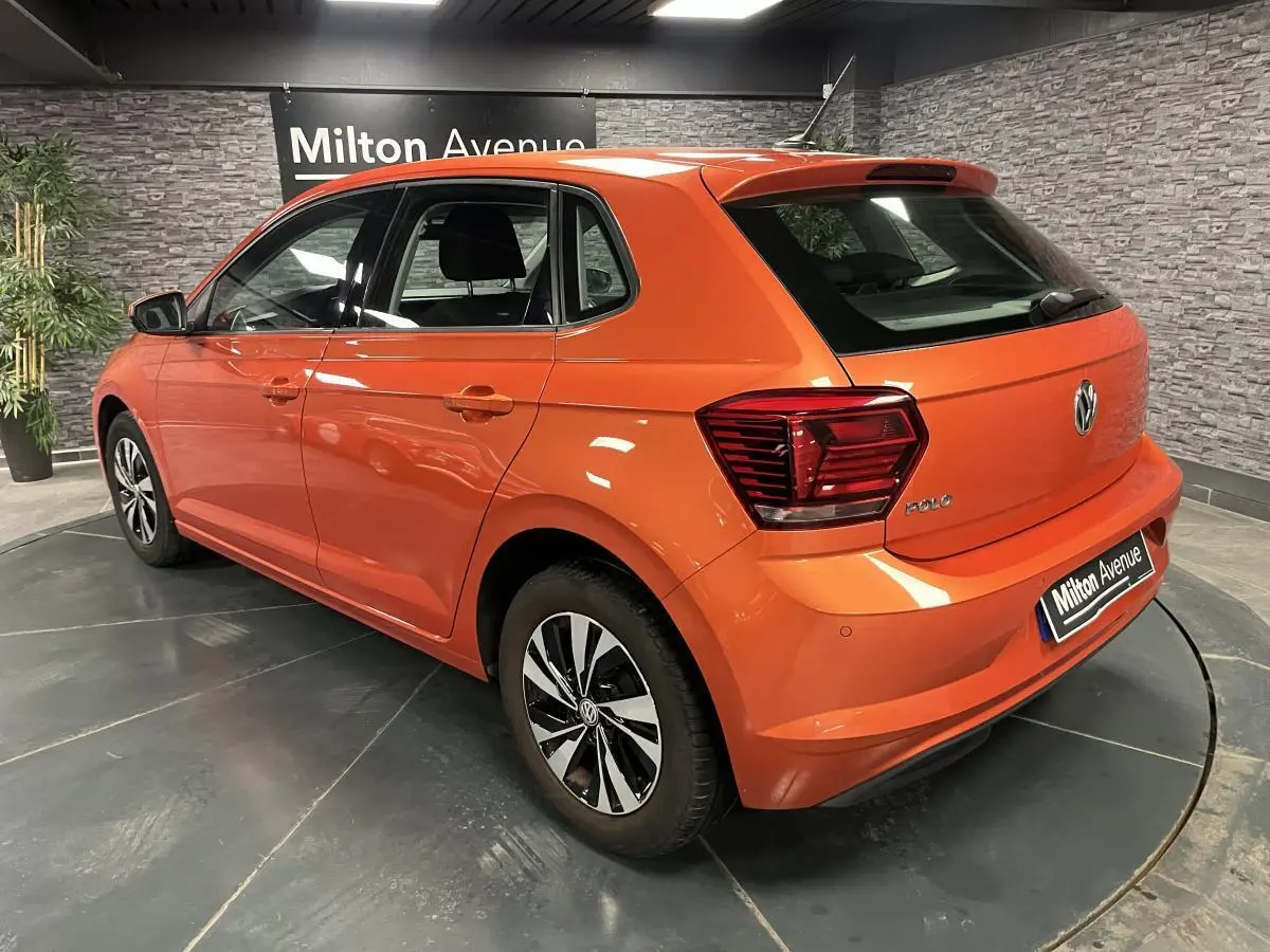 Vue 3/4 arrière droite d'une Volkswagen Polo orange métallisée avec jantes alliage et feux arrière LED dans un showroom.