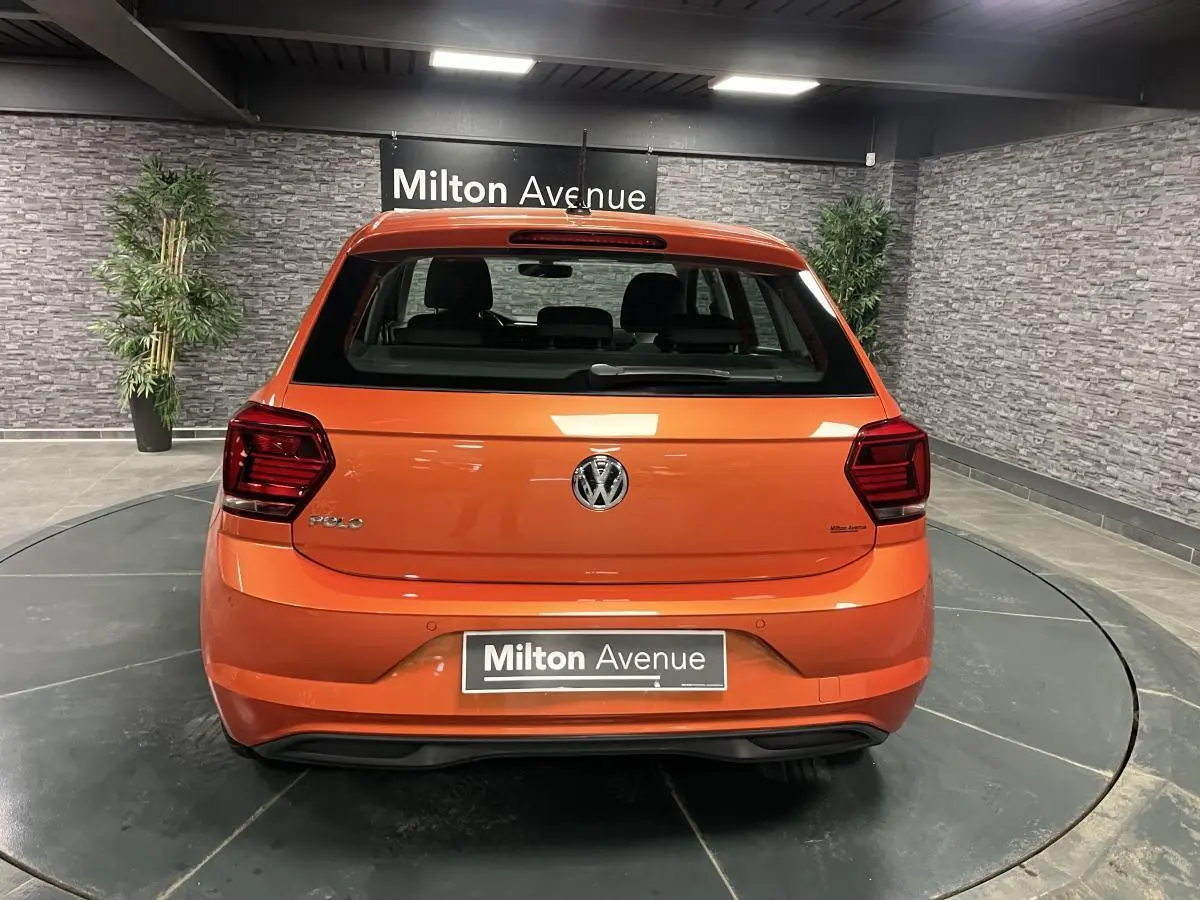 Vue arrière d'une Volkswagen Polo orange vif 2019, avec logo VW centré et feux arrière sombres en intérieur showroom.