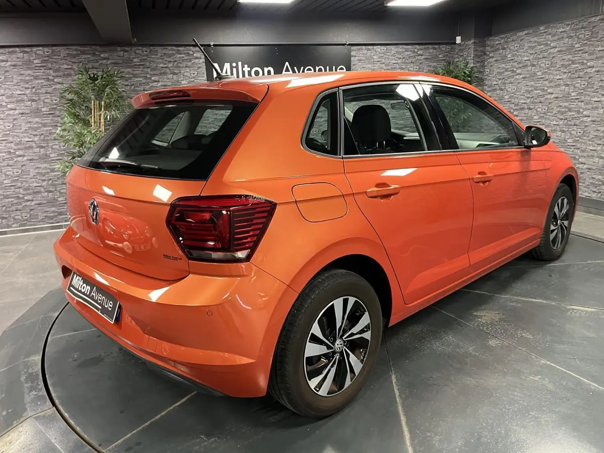 Volkswagen Polo orange vue 3/4 arrière droit, avec jantes alliage et feux arrière foncés en intérieur showroom.