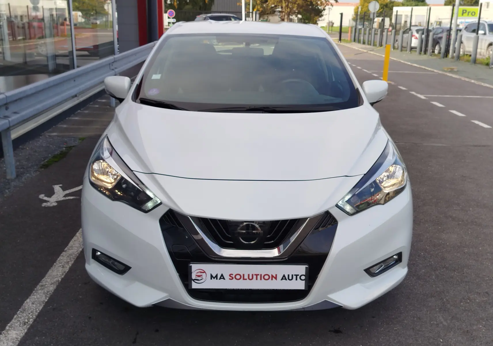 Vue avant d'une Nissan Micra blanche de 2019 avec phares allumés et calandre chromée sur parking urbain.