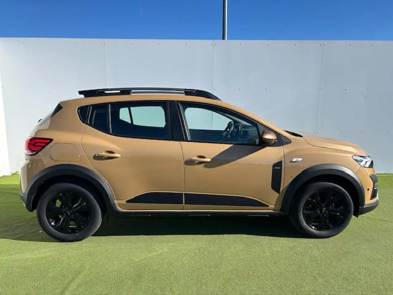 Profil droit d'une Dacia Sandero III Stepway beige Safari avec jantes noires et barres de toit gris Mégalithe.