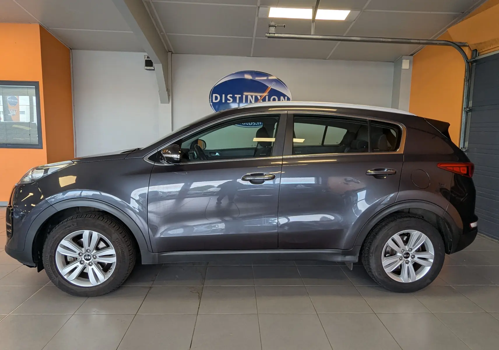 Vue de profil côté gauche d’un Kia Sportage gris 2016 en intérieur, avec jantes alliage et cinq portes visibles.