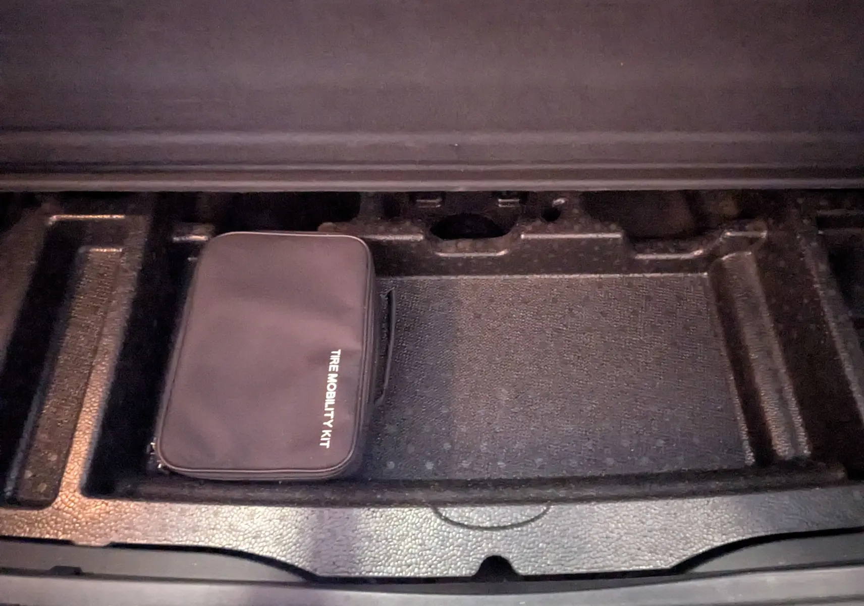 Compartiment de rangement noir dans le coffre d'un KIA Sportage gris, contenant un kit de mobilité pneus.