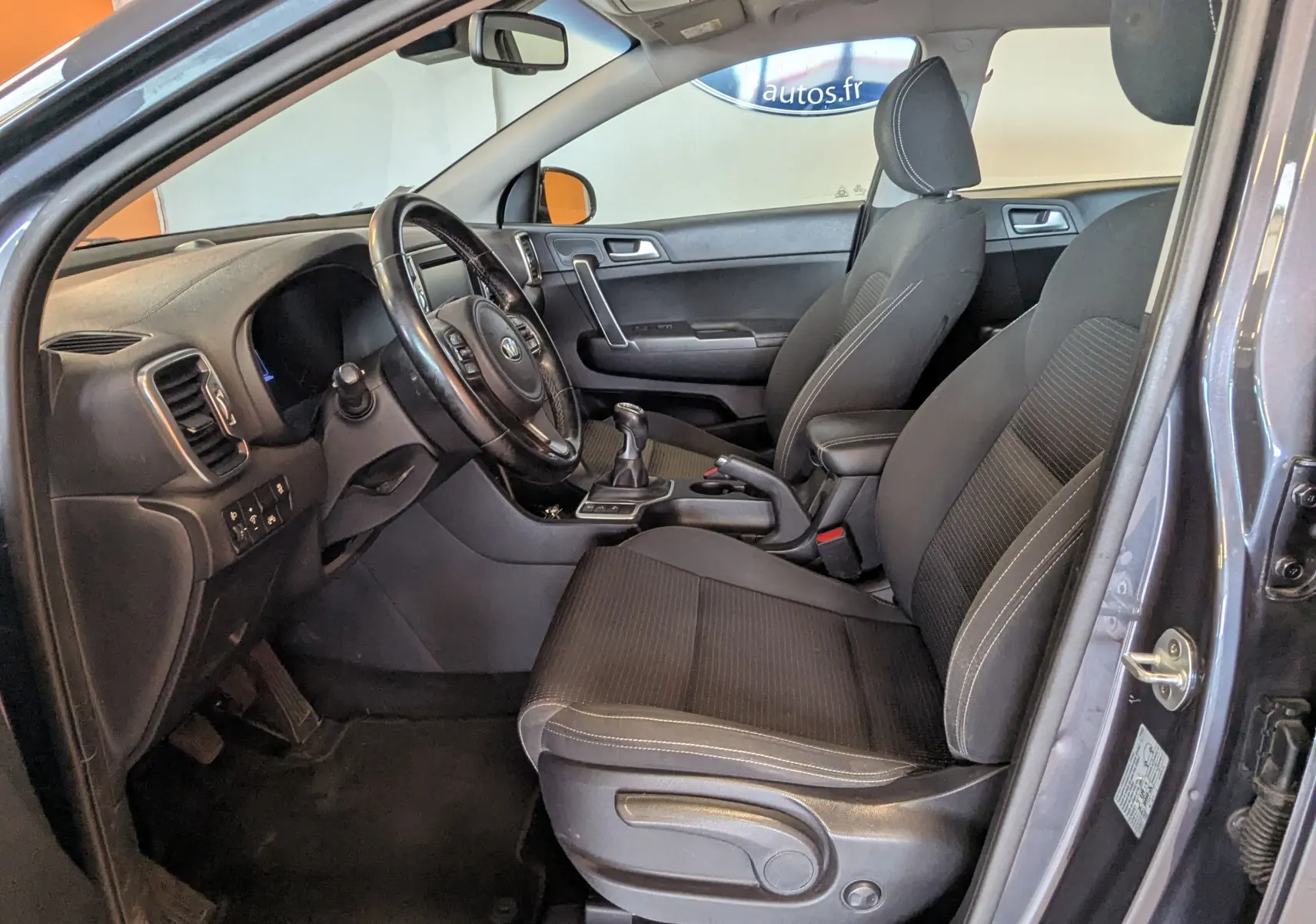 Vue intérieure côté conducteur du Kia Sportage gris 2016, montrant les sièges noirs et la console centrale avec levier de vitesses manuel.